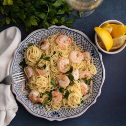Lay flat shot of Lemon Shrimp pasta plated on old world ceramic plate and lemons with white wine in a glass photographed on a dark blue background by SoftLight Photography