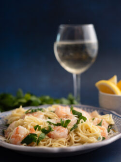 Lemon Shrimp pasta plated on old world ceramic plate and lemons with white wine in a glass photographed on a dark blue background by SoftLight Photography